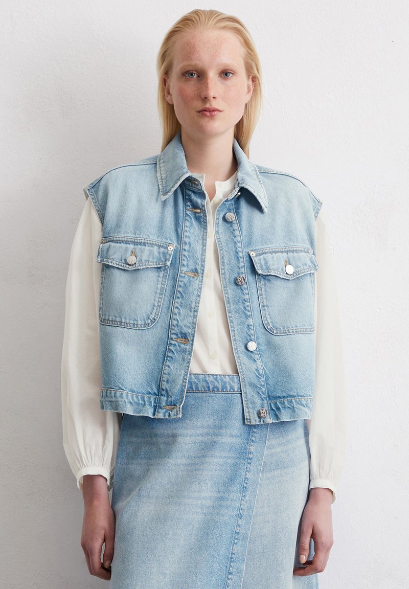 Leren spencer in lichtblauw met een kraag, twee voorzakken en zilveren hardware. G gedragen over een wit, langmouwshirt.