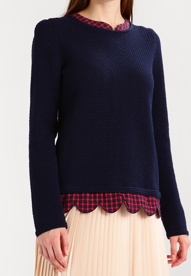 Femme portant un pull texturé bleu marine sur une chemise à carreaux rouge avec un ourlet festonné, associée à une jupe plissée beige clair.