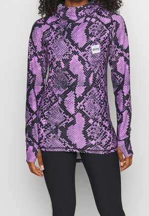 Kvinde iført en langærmet lilla og sort top med slangeprint og tommelhuller, kombineret med sorte leggings.