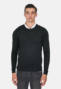 Maglione nero a maniche lunghe con scollo rotondo, realizzato in un tessuto a maglia liscia, con polsini e orlo a coste, indossato sopra una camicia bianca con colletto.
