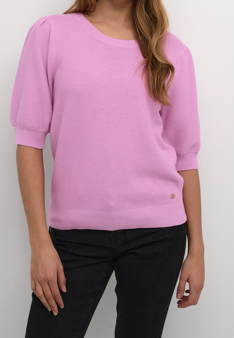 Pull en maille rose clair avec manches courtes bouffantes, poignets et ourlet côtelés, présentant un petit détail de logo près de la taille. Associé à un pantalon noir.