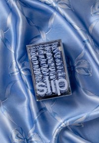 Hellblaue Seidenscrunchies in einer transparenten Box. Die Oberfläche ist glatt mit einer gerafften Textur. Der Hintergrund zeigt einen Satin-Stoff mit floralem Muster.