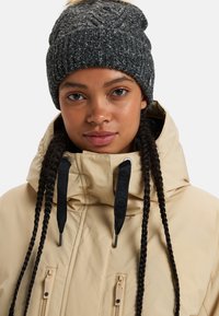 Giacca beige con colletto alto e cordini neri; abbinata a un berretto lavorato a maglia scuro con un pompon chiaro. Dettagli di design minimalista.