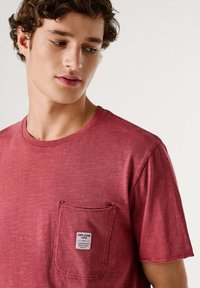 Camiseta de algodón roja con mangas cortas, escote redondo y un bolsillo en el pecho que presenta una etiqueta de ropa. Tejido texturizado con un acabado desgastado.