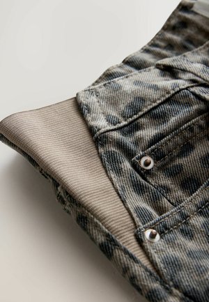 Denimkleidungsstück mit grauem und blauem Muster, ausgestattet mit einem beigen, gerippten Bündchen und metallischem Druckknopfdetail an der Tasche.
