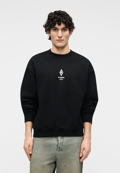 Νεαρός με σγουρά σκουροκάστανα μαλλιά, φοράει μαύρο φούτερ με λευκό λογότυπο και ανοιχτό γκρι τζιν, στέκεται μπροστά σε απλό φωτεινό φόντο.