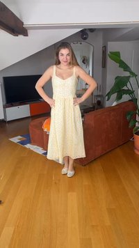 Ung kvinne i gul blomstrete kjole og flate sko står med hendene på hoftene i en moderne stue med tregulv og rustfarget sofa.