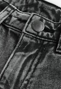 Gros plan sur un tissu en denim montrant un bouton métallique étiqueté "NEXT AUTHENTIC EST SINCE 1982" avec des coutures visibles.