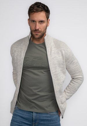 Lichtgrijze gebreide zip-up jas met geribde kraag, gelaagd over een olijfgroene t-shirt. Model geposeerd tegen een witte achtergrond.