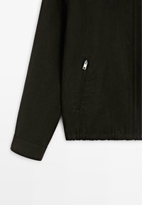 Manche et côté d'une veste noire avec une poche zippée et un ourlet élastiqué visibles sur un fond uni.