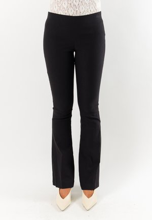 Donna che indossa pantaloni a zampa neri aderenti e tacchi bianchi a punta su sfondo bianco semplice.