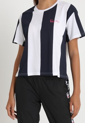 Koszulka z bawełny z pionowymi granatowo-białymi paskami, z różowym logo na piersi i luźnym krojem. Widoczne czarne spodnie sportowe.