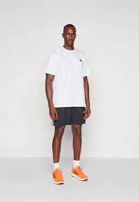 Vit atletisk tröja med korta ärmar och svart logotyp, kombinerad med svarta shorts med vita ränder, och orangea träningsskor med svarta detaljer.