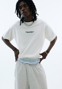 White short-sleeve t-shirt with blue embroidered text. Paired with light gray sweatpants featuring a blue-striped waistband.