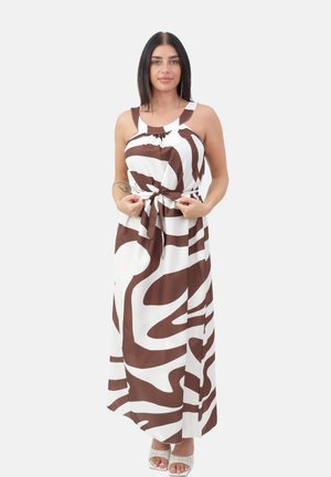 Kvinde med langt mørkt hår iført en hvid og brun maxikjole med abstrakt mønster uden ærmer, justerer taljebåndet, står på hvid baggrund.