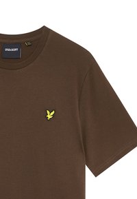 T-shirt in cotone marrone con scollatura rotonda. Logo giallo ricamato ben visibile sul lato sinistro del petto. Texture liscia, maniche corte.