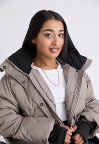 Puffy beige jacket with a black collar, featuring snap buttons and elastic cuffs, layered over a white top with a gold necklace.