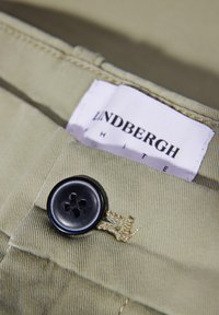 Helles olivgrünes Gewebe mit einer glatten Textur, das einen großen schwarzen Knopf und ein weißes Etikett mit dem Text "Lindbergh White" aufweist.