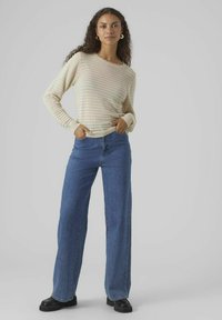 Maglione a maniche lunghe color crema, lavorato a maglia con un motivo testurizzato, abbinato a jeans blu a vita alta e a gamba larga e scarpe con tacco in nero.