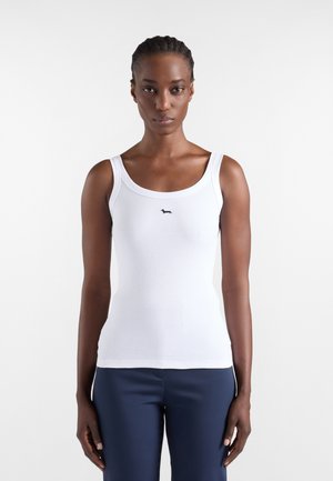 Nainen, jolla on letitetyt hiukset ja valkoinen ribbineulottu toppi, jossa on pieni musta koiramerkki, yhdistetty tummansinisiin housuihin, seisoo valkoista taustaa vasten.