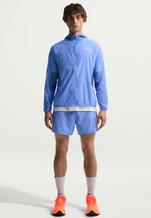 Jonge man staat gekleed in een lichtblauw sportjack en korte broek, witte sokken en feloranje hardloopschoenen tegen een effen achtergrond.