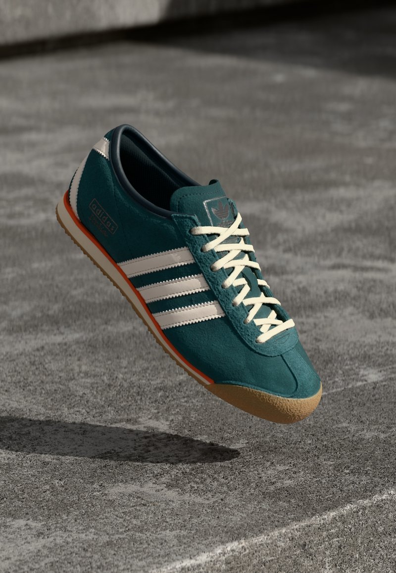 Sapatilhas de camurça verde-água com riscas brancas e destaques em laranja. Apresenta uma sola de borracha texturizada e atacadores de cor clara. Design atlético clássico.