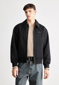 Sort bomberjakke med en tekstureret overflade, præget krave og lynlås, båret over en beige strikket sweater og blå jeans.