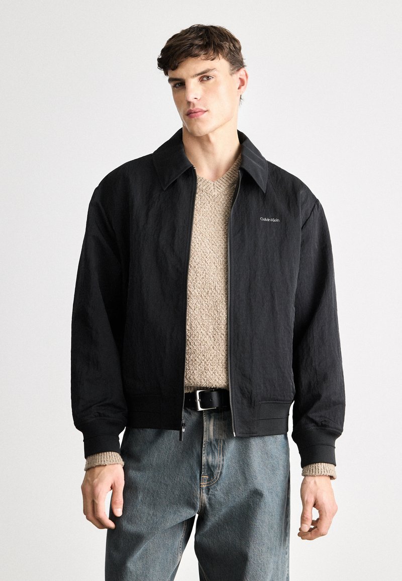 Sort bomberjakke med en tekstureret overflade, præget krave og lynlås, båret over en beige strikket sweater og blå jeans.