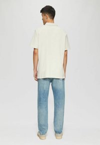 Hellbeige Kurzarm-Polohemd mit Kragen, kombiniert mit hellblauen Jeans und beigen Schuhen, von hinten betrachtet vor einem neutralen Hintergrund.