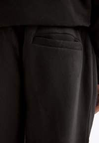 Zwarte sweatpants met een gladde textuur hebben een achterzak met een horizontale opening, ontworpen voor comfort en functionaliteit.