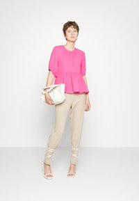 Top peplum fúcsia com decote franzido, combinado com calças beje de corte elegante com detalhes de cordões. Modelo segura uma mala branca.