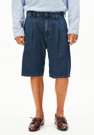 Mann trägt dunkelblaue kniehohe Denim-Shorts, hellblaues langärmeliges Hemd und braune Schnürschuhe aus Leder und steht.