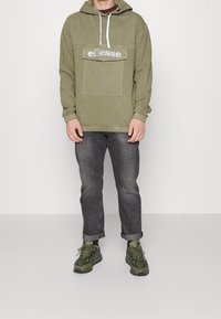 Sudadera verde oliva con logo en el bolsillo delantero, capucha con cordón y puños elásticos. Combinada con jeans gris oscuro y zapatillas verde oliva.