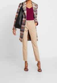 Karierter langer Mantel in Beige, Braun und Marineblau, kombiniert mit einem burgunderfarbenen Oberteil und beigen engen Hosen. Eine schwarze Tote-Tasche und Riemchensandalen runden den Look ab.