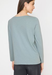 Maglietta a maniche lunghe in morbido tessuto blu chiaro con scollatura ampia e superficie liscia. Esposta da dietro, che evidenzia i dettagli delle cuciture.