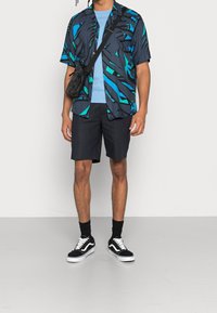 Lyhythihainen nappisuljettava paitapusero, jossa on laivastonsininen ja teevihreä abstrakti kuvio, yhdistettynä vaaleansiniseen t-paitaan, laivastonsinisiin shortseihin, mustiin sukkiin ja mustiin lenkkareihin.