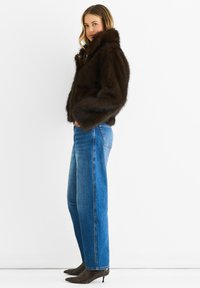 Woman wearing a dark brown fur jacket, blue high-waisted jeans, and pointed-toe ankle boots with a low heel, standing against white background.