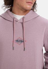 Sudadera de color púrpura claro con cordones, que presenta un logotipo de diamante en negro y rojo en el pecho. Textura suave, diseño casual.