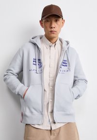 Svetlo šedá mikina na zips s prednými vreckami, zdobená fialovým logom. Nosená cez béžovú košeľu na gombíky, doplnená hnedou čiapkou.