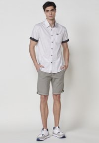 Camisa de botones de manga corta en gris claro con un bolsillo en el pecho y ribete oscuro, combinada con pantalones cortos caqui enrollados y zapatillas blancas.