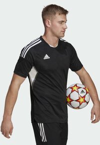 Black athletic jersey with white stripes, textured circular pattern, and Adidas logo. Model holding a white soccer ball with red and yellow stars.