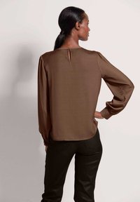 Braune Satinbluse mit langen Ärmeln und einem Schlüsselaugen-Detail im Rücken, kombiniert mit schwarzen Hosen. Glatte Textur und lockere Passform.