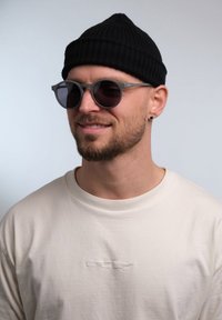 Occhiali da sole grigi con lenti rotonde, una montatura sottile e un logo impresso. Indossati con una camicia beige chiaro e un berretto in maglia nero.