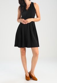 Black sleeveless dress with a V-neckline, flared skirt, and textured fabric. Worn with tan suede heels. Simple design and clean lines.