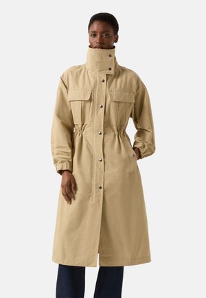 Kvinde iført lang beige frakke med høj krave, knaplukning, lommer med klap foran, indsnævret talje og elastiske manchetter, stående mod ensfarvet baggrund.