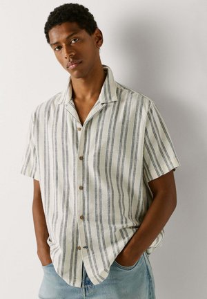 Nuori mies, jolla on lyhythihainen valkoinen paita, jossa on pystysuorat mustat raidat, ja vaaleansiniset farkut, seisoo kädet taskuissa yksinkertaisen seinän edessä.