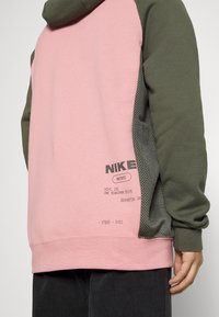 Person som bär en långärmad hoodie i rosa och olivgrön med svarta mesh-sidopaneler och svart text nära fållen på vänster baksidan.