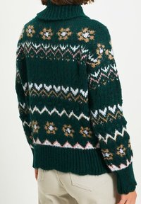 Henkilö, joka pukeutuu tummanvihreään neulotuun villapaitaan, jossa on monivärisiä sawtulaitteita ja kukkakuvioita, yhdistettynä vaaleanbeigeihin housuihin, selkä kameraan päin.