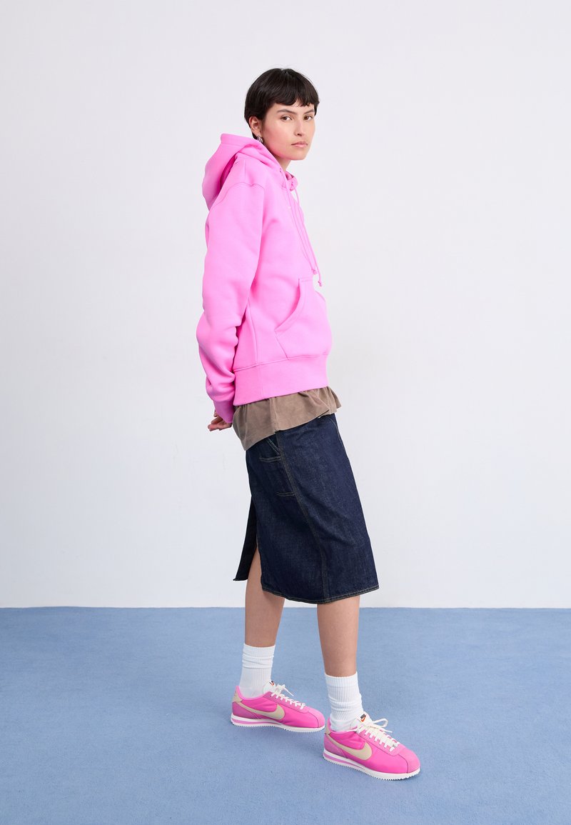 Helles, pinkes Hoodie mit einer Vorderasche, über einem braunen Shirt getragen; kombiniert mit einem knielangen Jeansrock und rosa Nike-Sneakern.