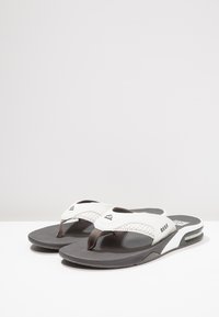 Des tongs blanches et grises avec une semelle intérieure texturée, une semelle en caoutchouc et des sangles réglables avec un détail de logo.